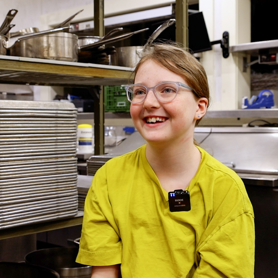 A young student sits in a professional kitchen, speaking to a camera.