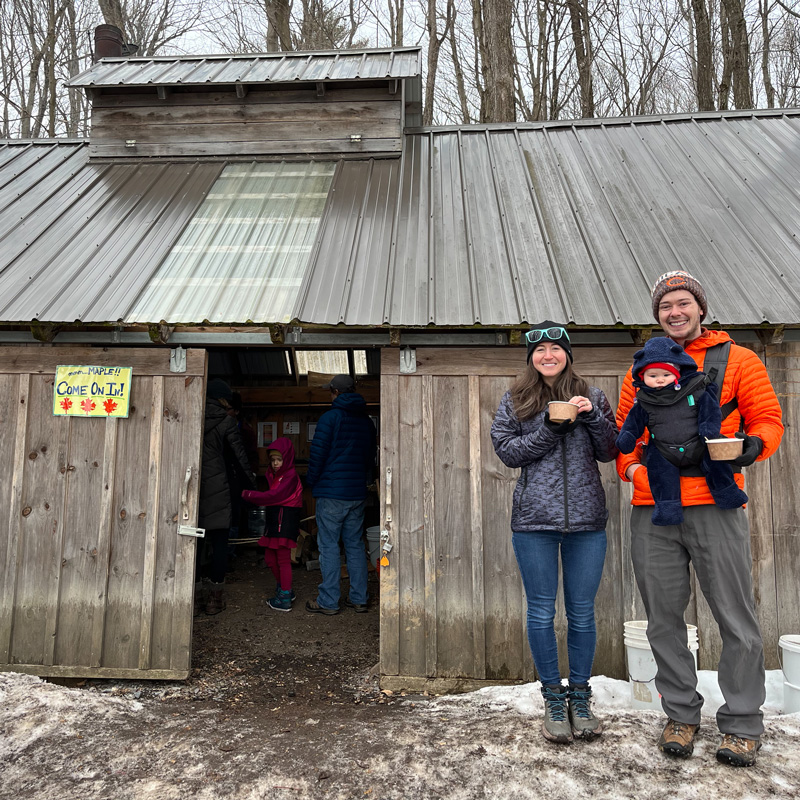  Generate by AI Assist  Two adults and a baby standing in front of a rustic sugar shack with a sign reading "Come on In!". They are dressed in winter clothing and smiling.