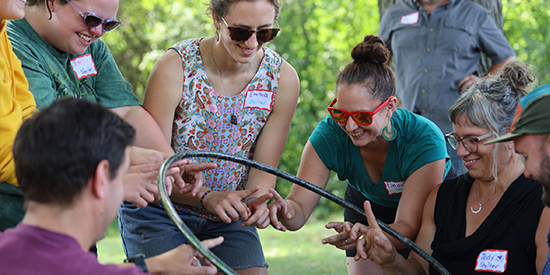 Seven adults smile while balancing a hula hoops on their hands in an outdoor team building activity