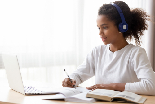 African American teen girl wearing headphones learning language online_ using laptop_ looking at screen_ doing school tasks at home_ writing notes_ listening to lecture or music_ distance education
