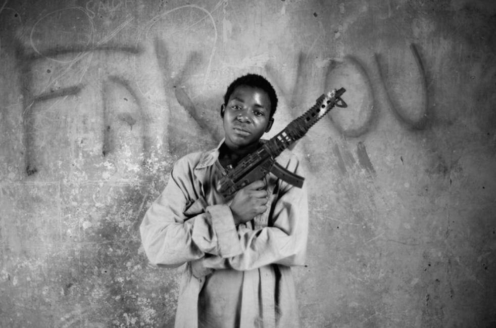 A child holding a toy machine gun stands in front of graffiti written on an urban cement wall
