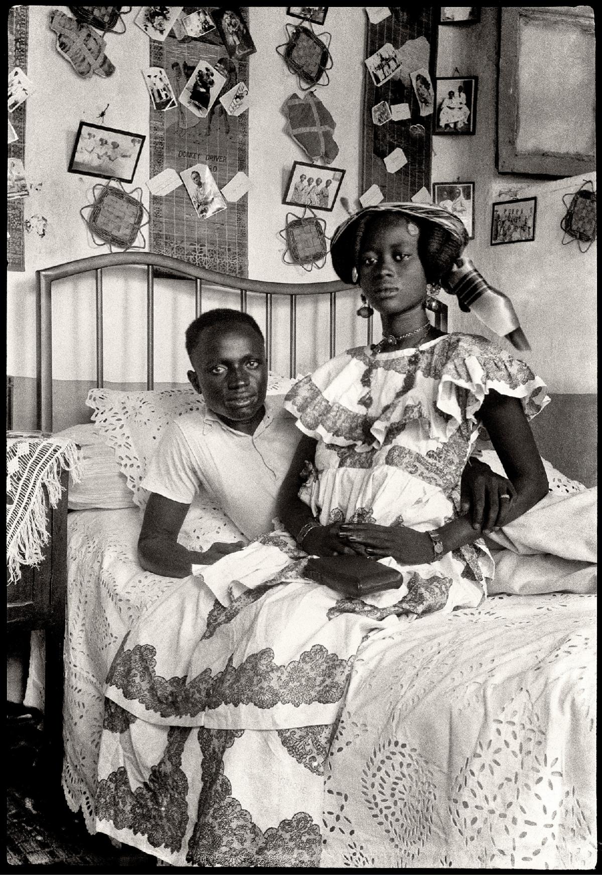 A man and a woman sitting on a bed surrounded by photographs on the walls