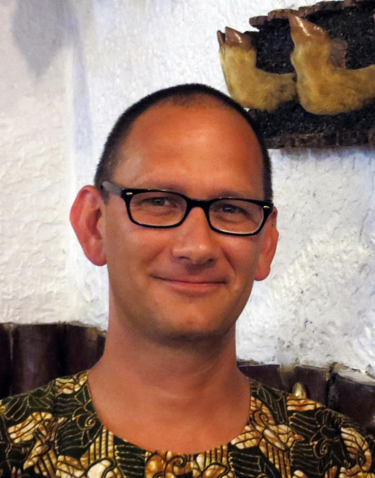 A headshot photograph of a man with black rimmed glasses and a patterned shirt