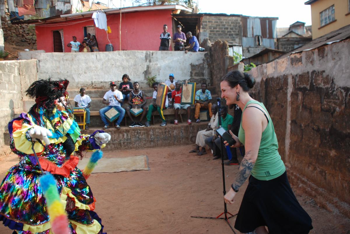 A masked performer in brightly colored textile ensemble dances with a smiling woman in a green shirt.
