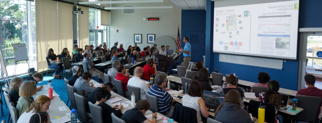 A scientist speaks to a room full of students at tables
