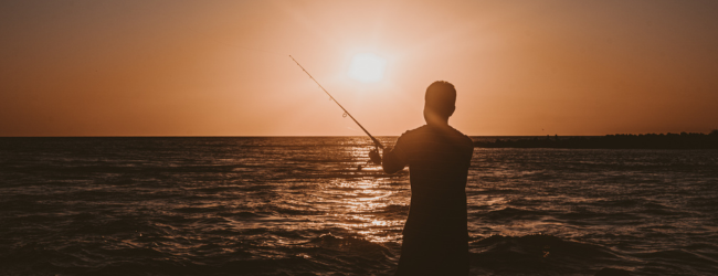 A person fishing on the shore of the ocean at sunset