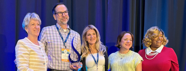 Five people stand with a blue curtain behind them. One holds an award.