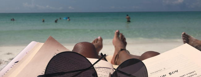 A view of the ocean at the beach over a person's bottom half with a book and sunglasses