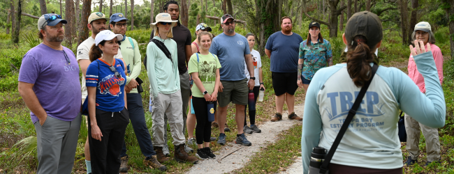 Someone in a TBEP shirt talks to a crowd in a forested setting