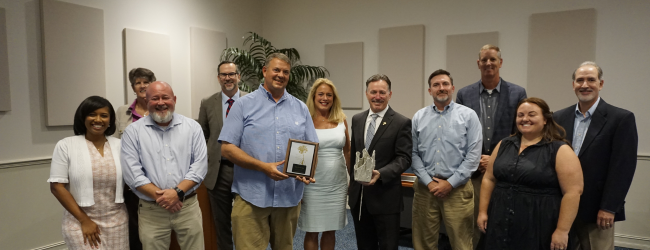 Eleven people stand in a meeting room, one holds an award, another holds an oyster rag pot