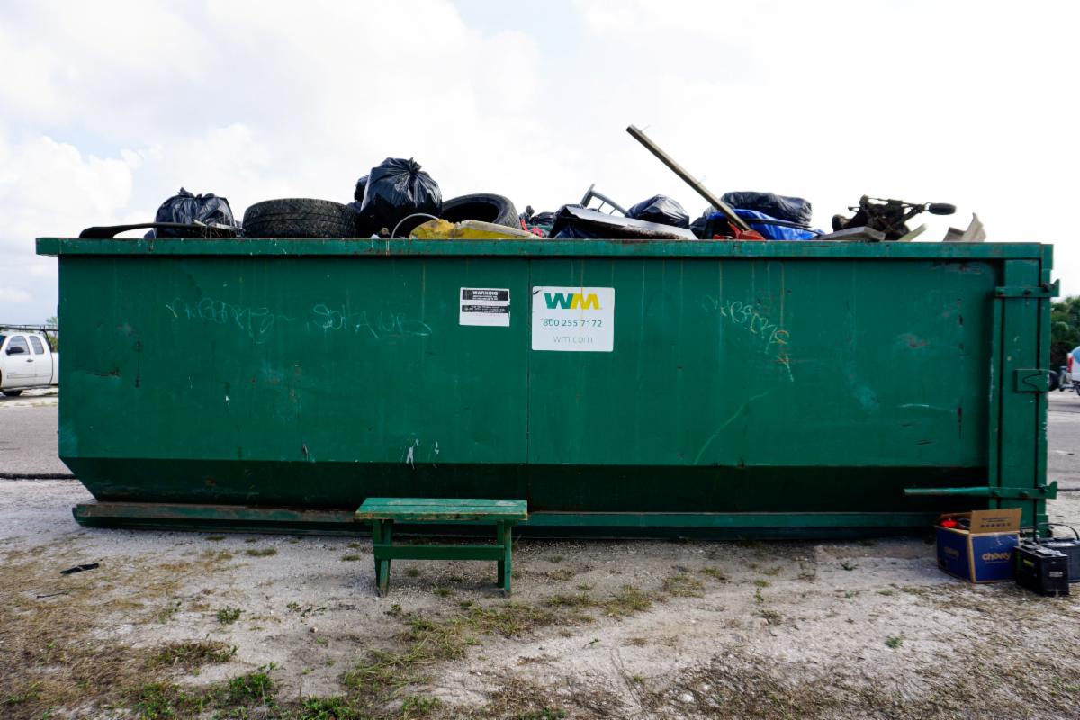 A large green dumpster is overflowing with garbage bags and debris