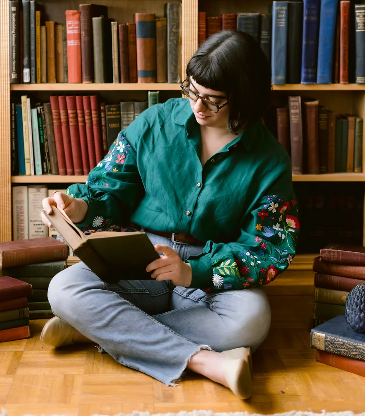 Lizzy Saxe is seen in a portrait style photo sitting on a floor in front of a bookcase reading a book. 