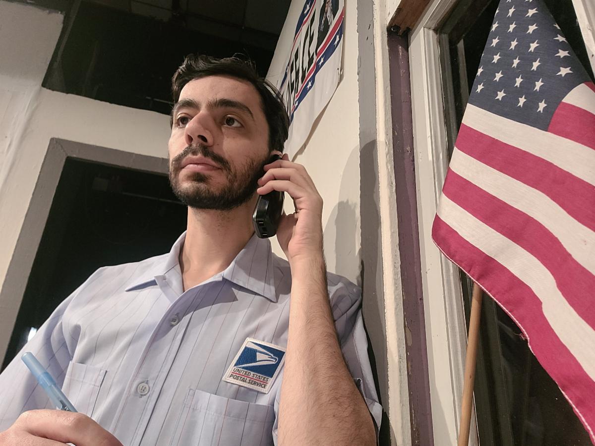 Emanuel is posing talking on a phone wearing a post office uniform and standing in front of an American flag.