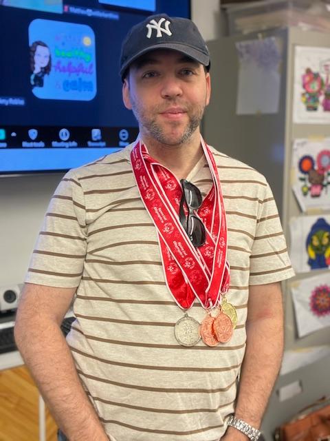 Kevin stands wearing a striped shirt a ballcap and his medals on his chest. 