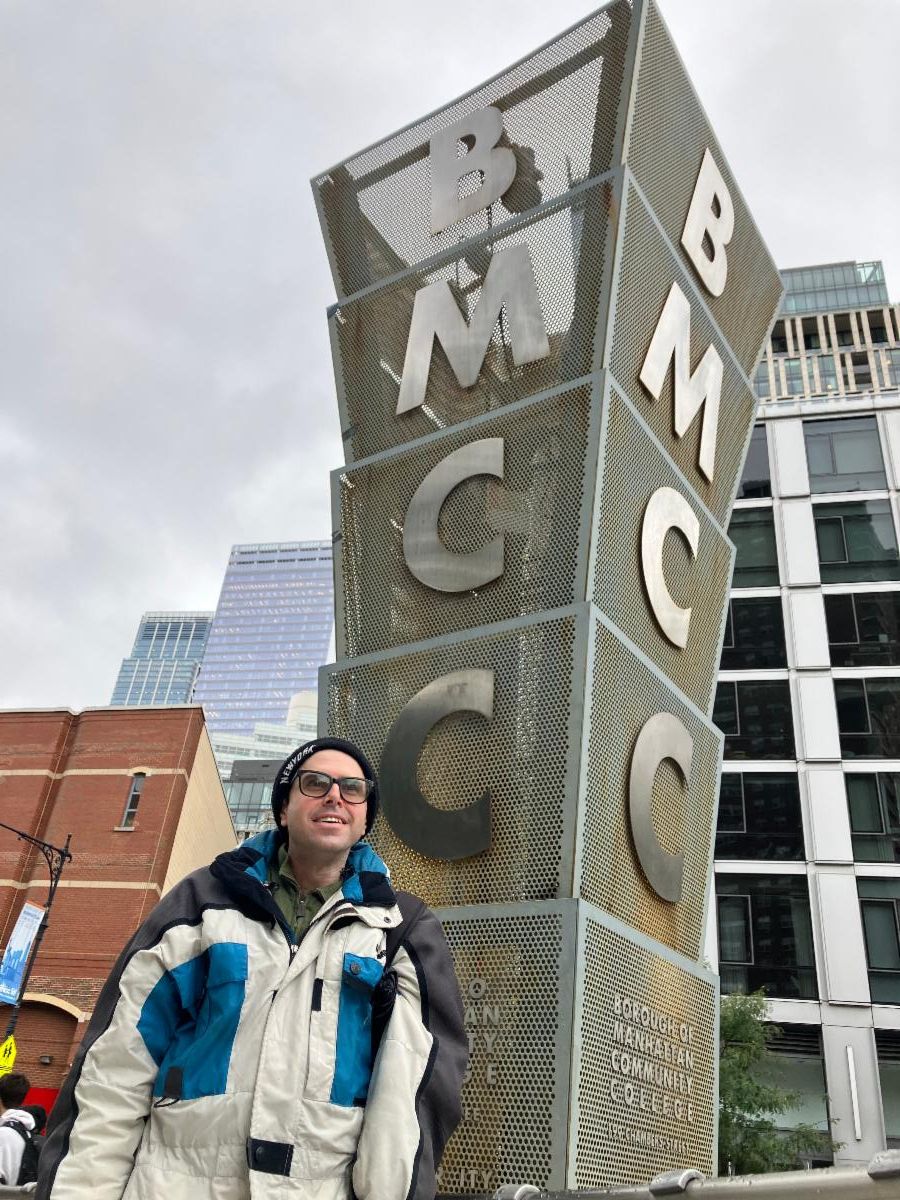 Rusty stands in front of the BMCC sign outside campus wearing a warm coat. 