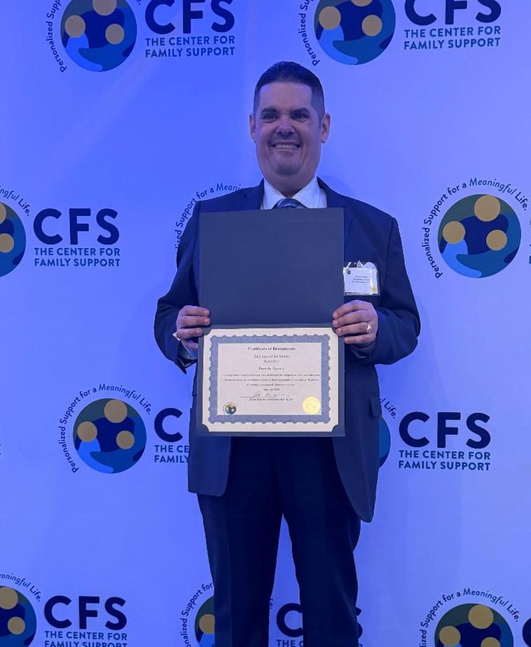 Ernesto stands holding a certificate in front of a screen with the CFS logo. The lighting  has a blue tint and he his wearing a collared shirt and a big smile. 