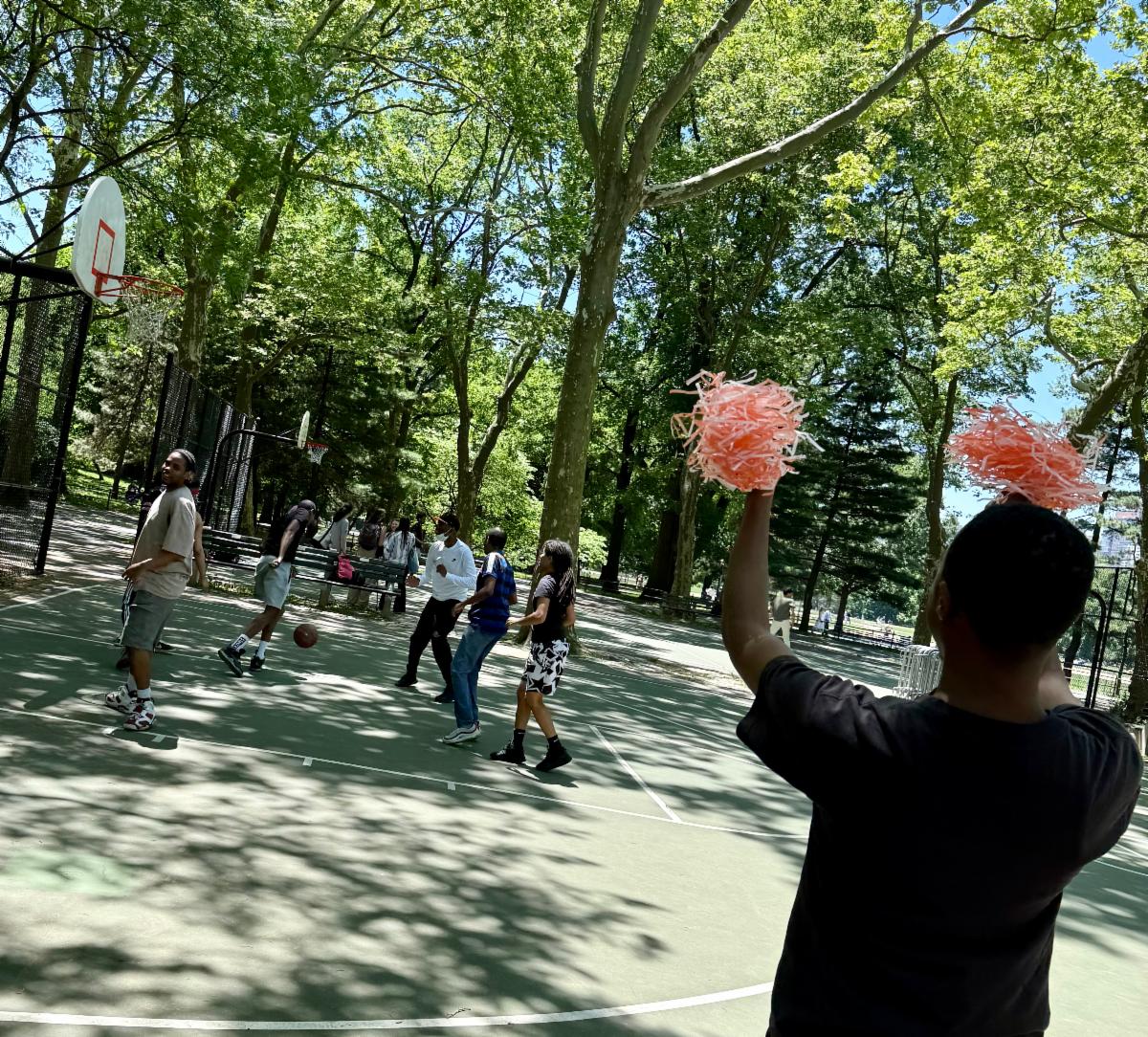 Seen from behind, a person waves orange and white pom-poms while others play basketball. 