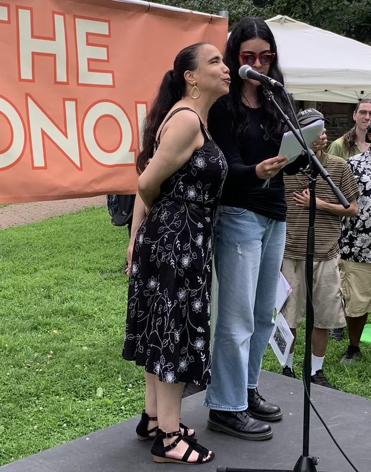 Noemi stands at a microphone wearing a long black sundress with a pink banner sign behind her.