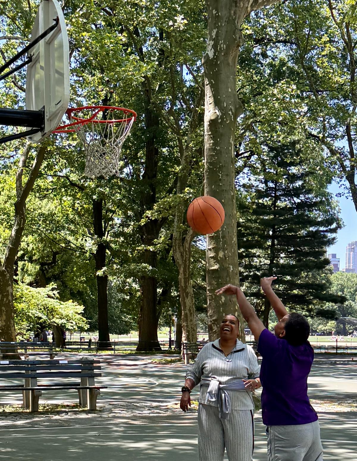 Two people on a sun-dappled basketball court - one takes a shot and the other is smiling in encouragement. 