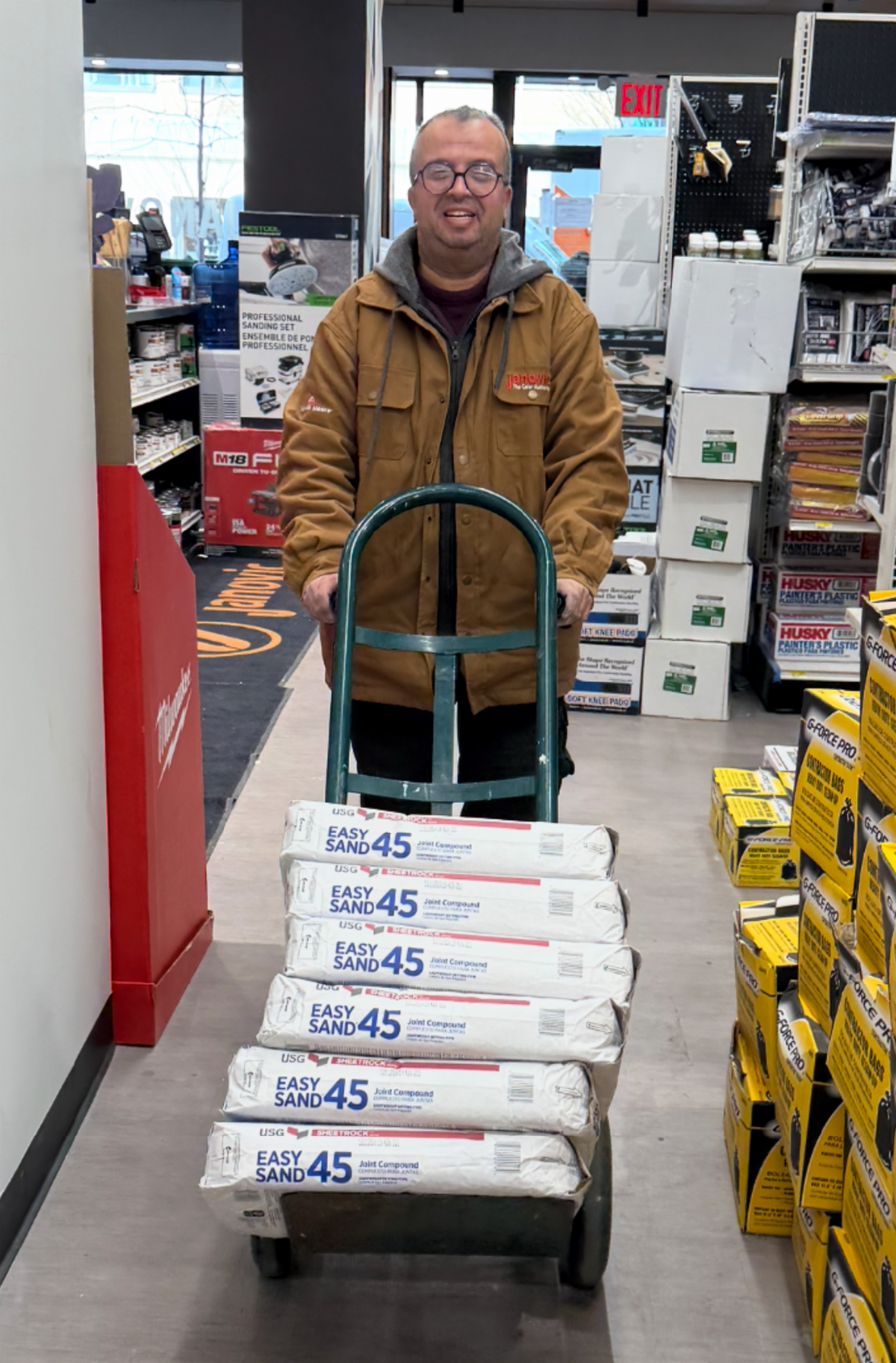 Zeiad stands wearing a heavy coat that has the Janovic logo on it and he's pushing a hand truck that has heavy sacks on top of it. 