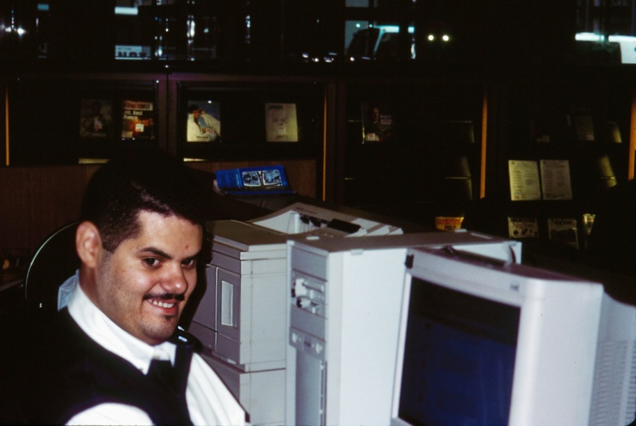 Ernesto is seen in what is clearly an older photograph wearing a mustache and sitting in front of an old boxy desk top computer. 