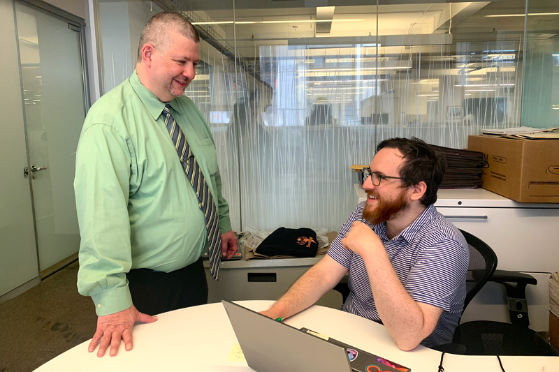 Jim is wearing a light green shirt and standing and Ben wears a purple shirt and is sitting at a table. Both are in an office. 