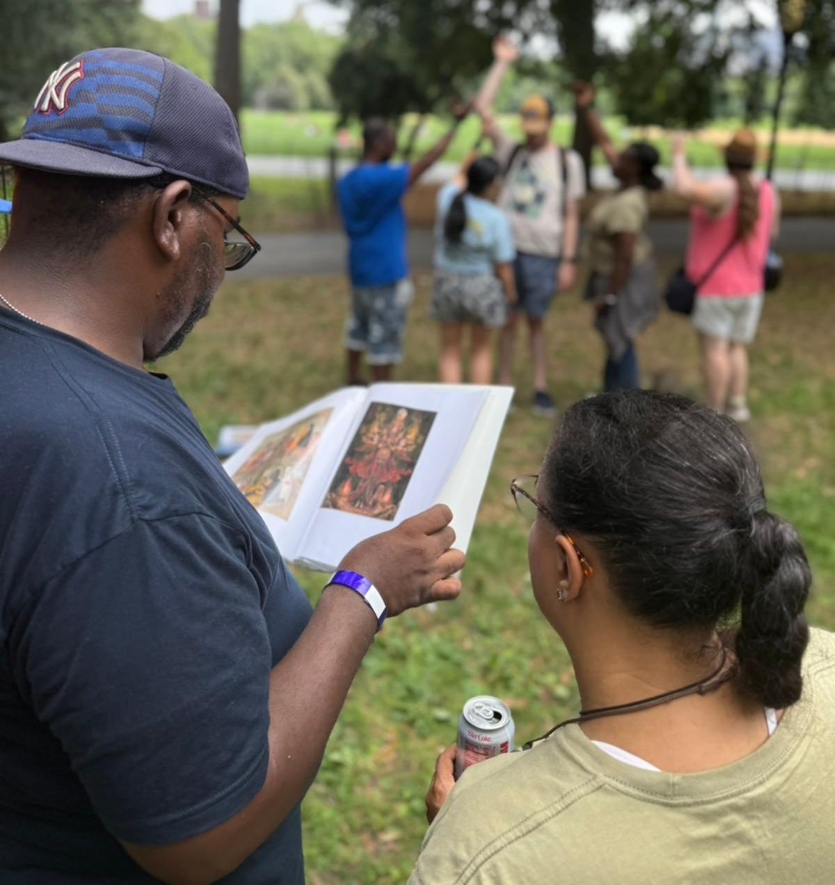 Two people study an images in a book, while in the background a group of people are standing in a unique formation. 