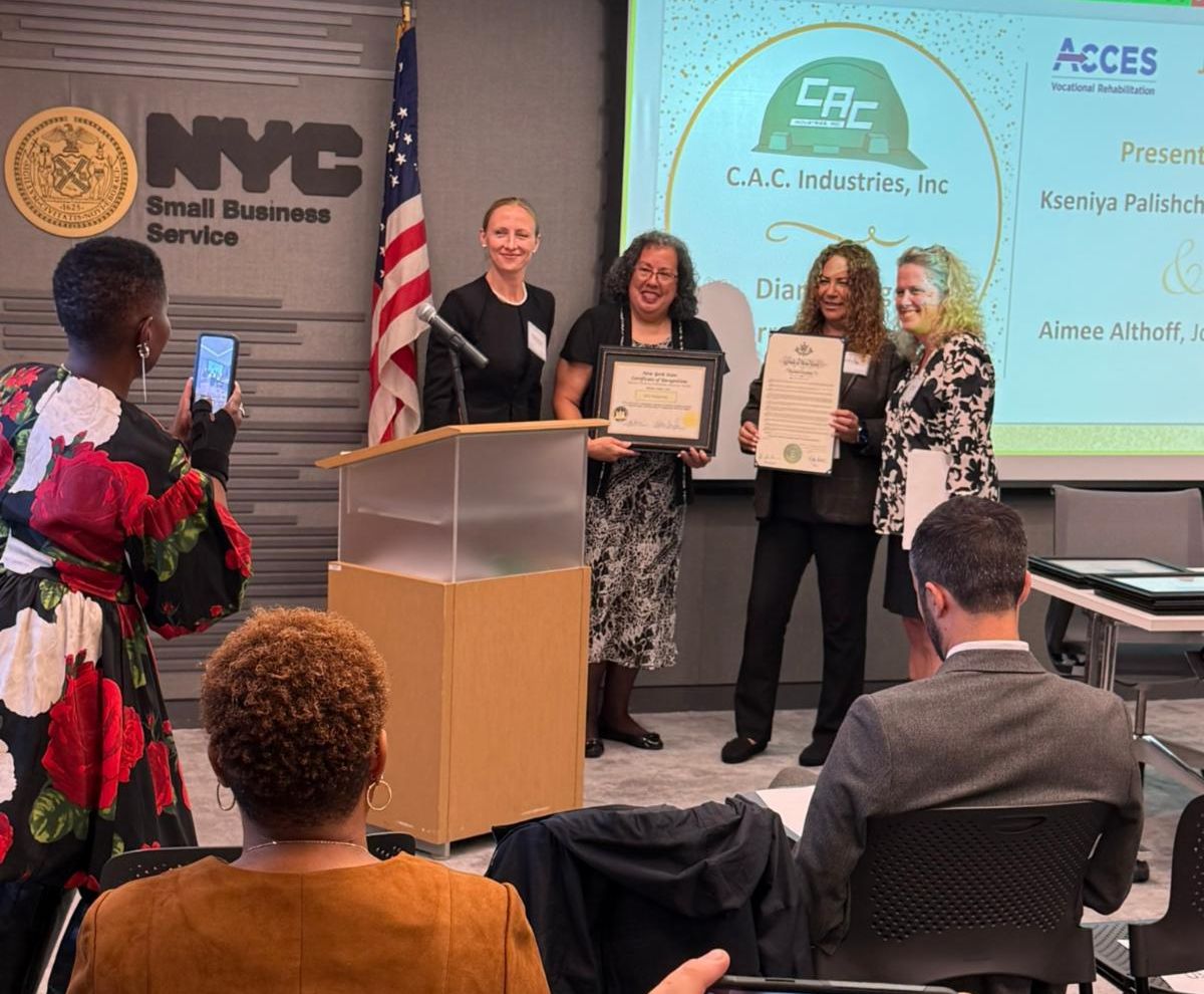 A group of people are standing in front of a conference room audience holding an award in front of a flag.
