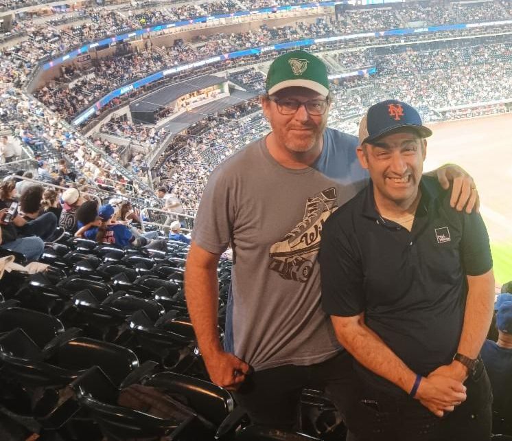 Josh and Rob are in the stands of the ballfield with the playing field behind them. 