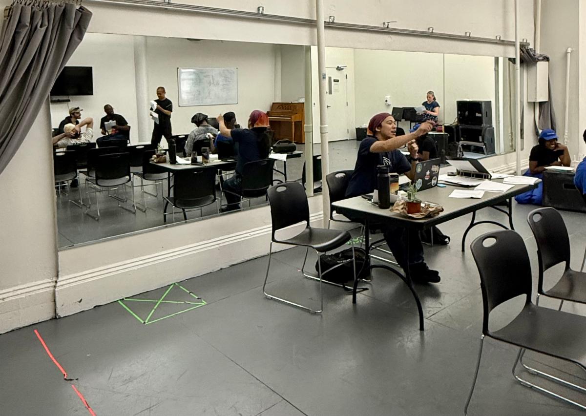 Anson is seen sitting at a table giving a directive. Behind him is a mirror that reflects several members of the cast of the improv show after just finishing a scene.