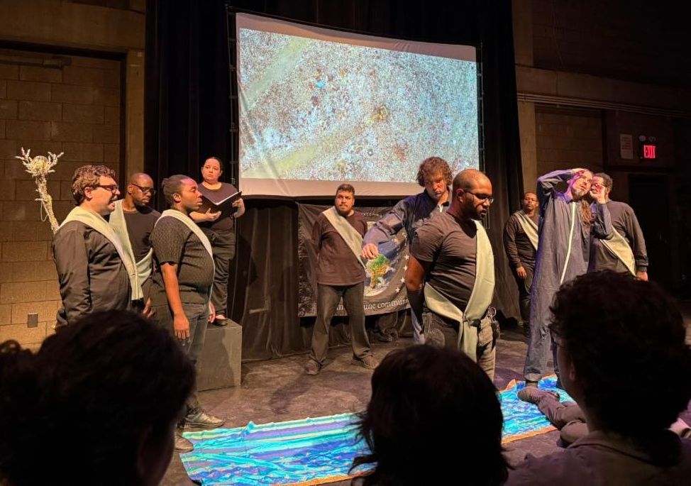 The cast is on stage during a scene with a screen behind them showing various images.