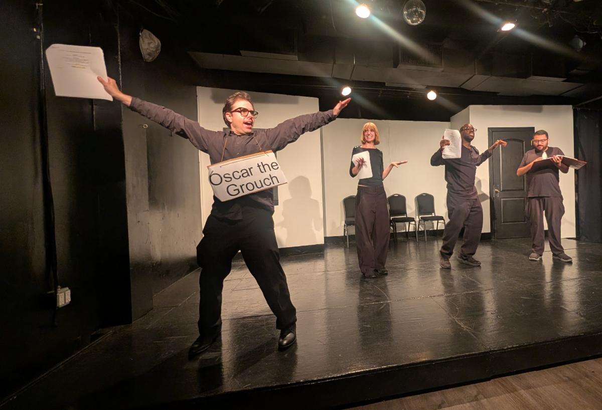 Performers wearing black hit various dramatic poses, wearing black on a black and white stage. One wears an Oscar the Grouch sign.