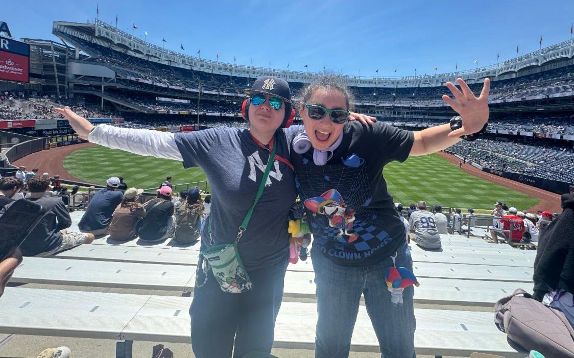Two people stand in front of a wide shot of Yankee stadium, wearing sunglasses. 