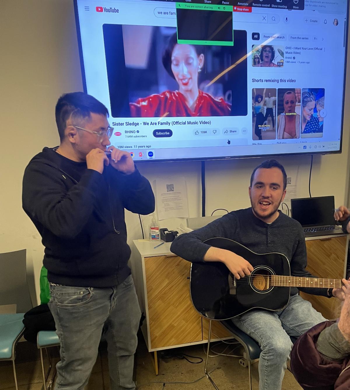 Max sits with a guitar and Camden is standing beside him in the Job Path conference room. On the screen near them is a YouTube video playing Sister Sledge "We Are Family."