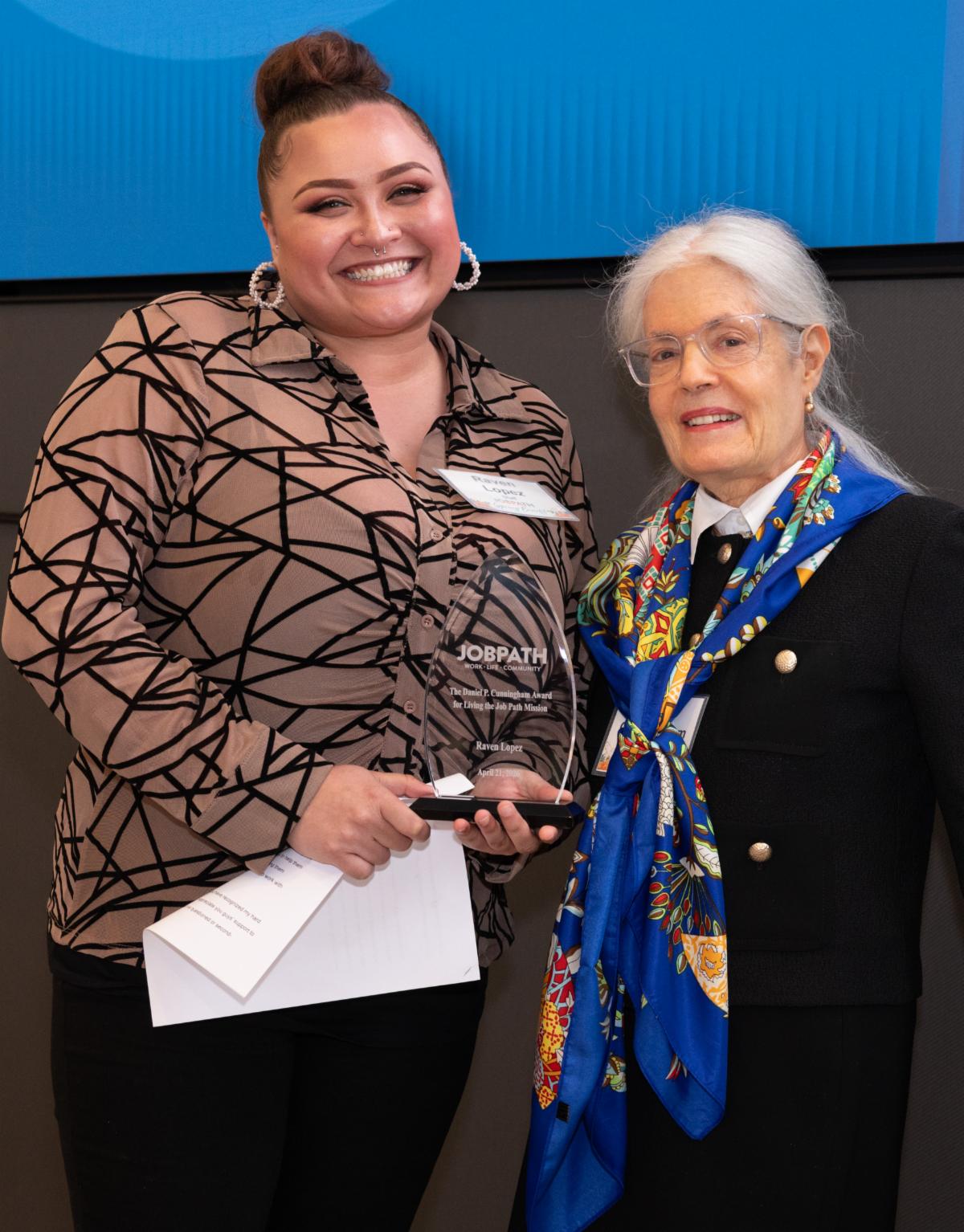 Raven and Alice smile while holding a glass award in front of a blue background. 