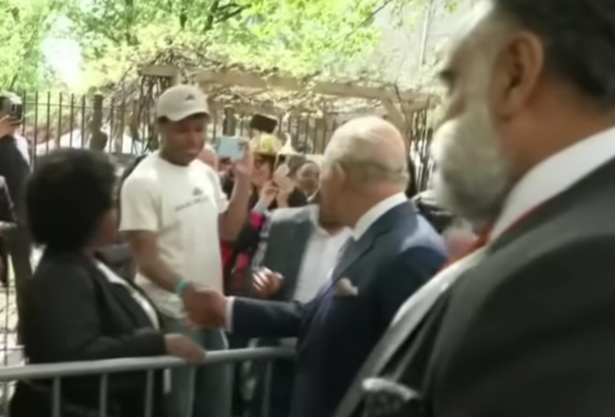 A slightly blurry still photo shows Chris shaking hands with England's King Charles III. 