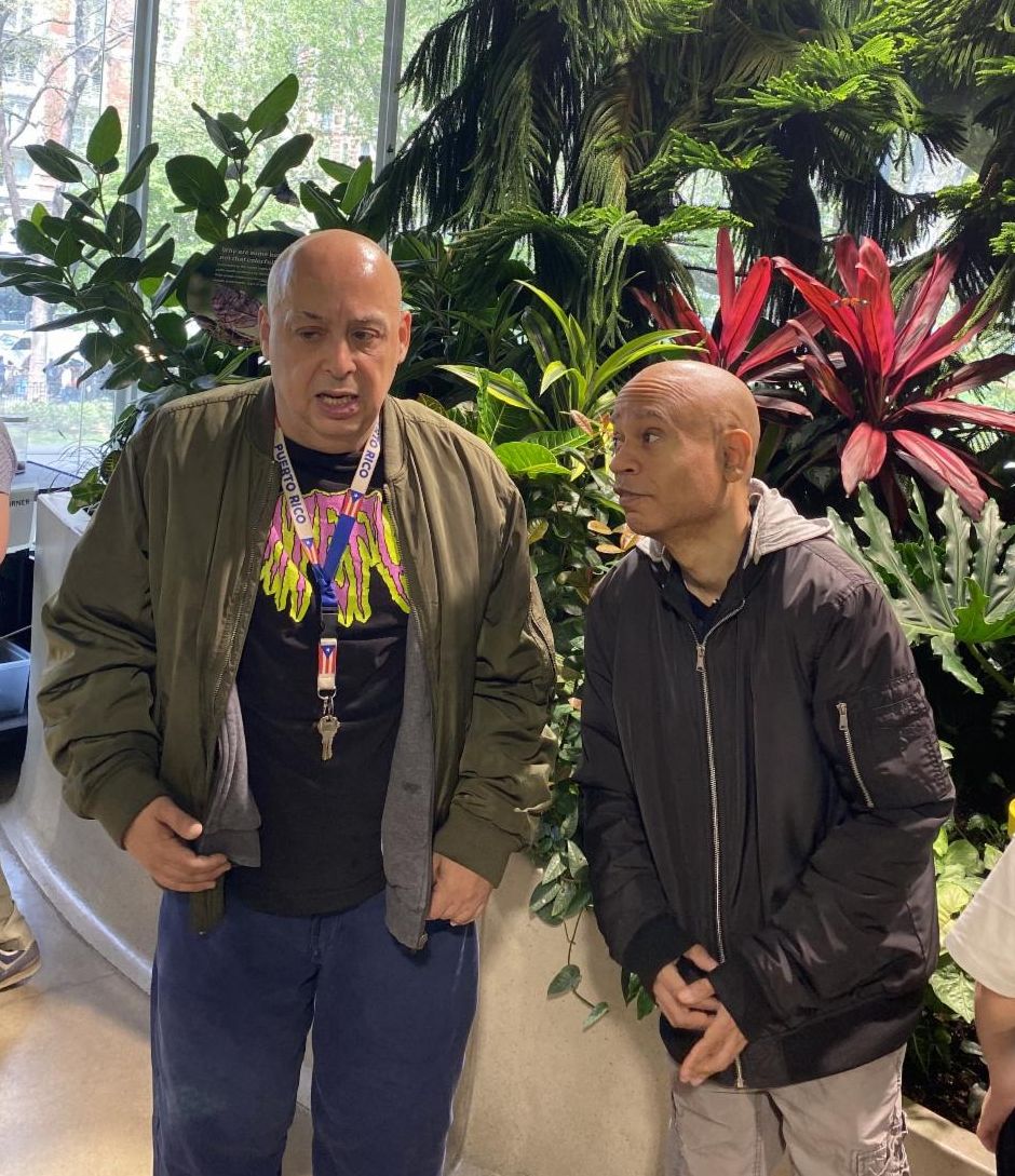 Cornell and Steven are in a lush tropical room with plants and flowers. 