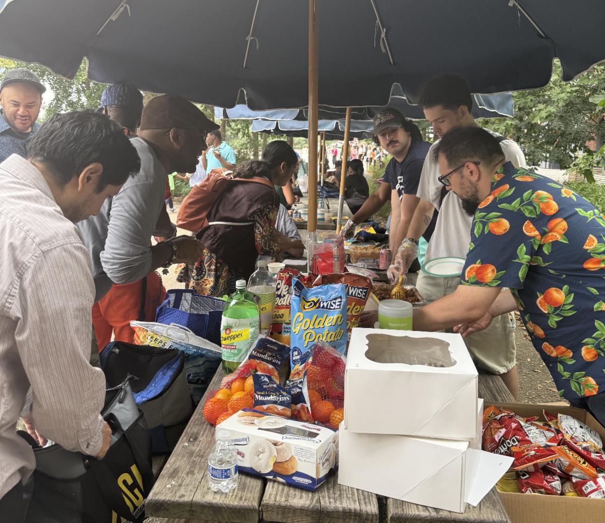 People serving and receiving food under a picnic table umbrella