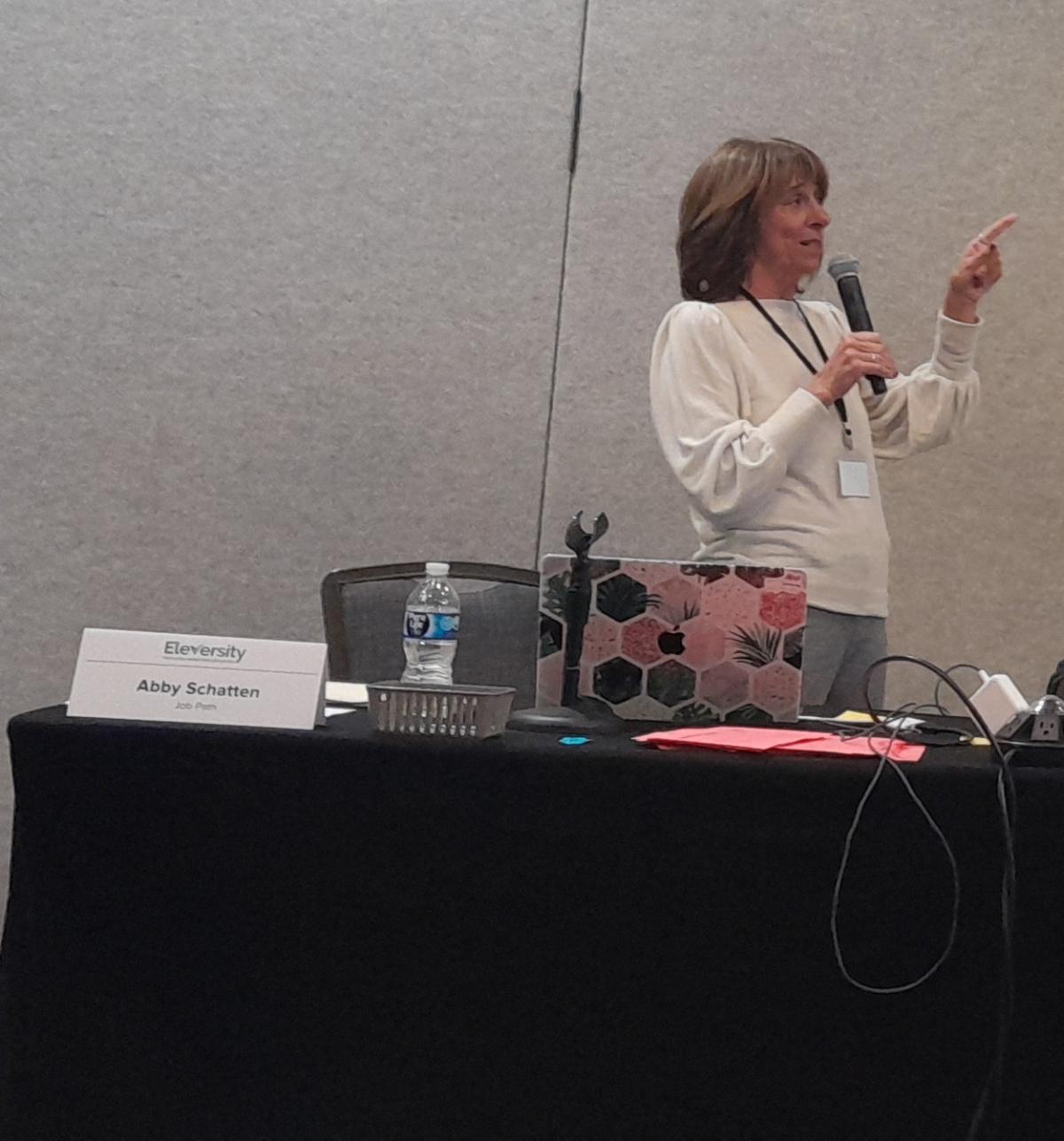 Abby is standing at a table and gesturing. 