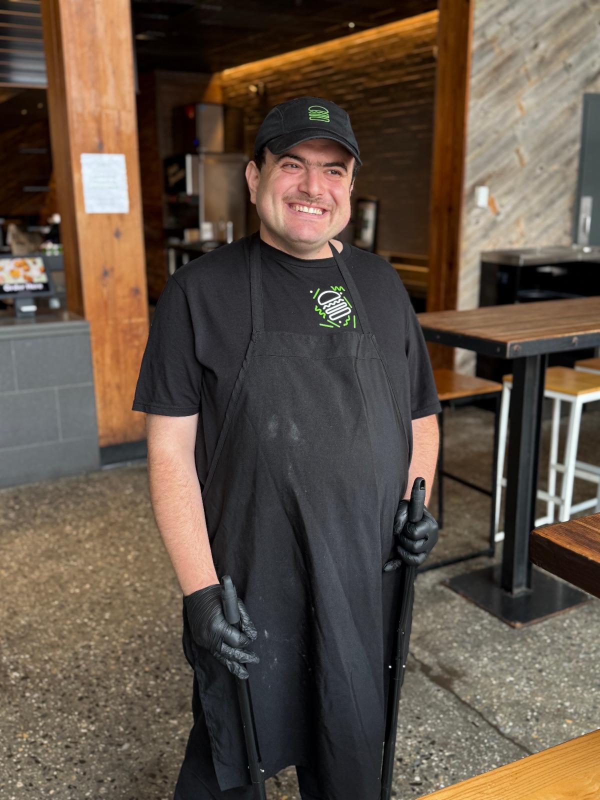 Oscar stands wearing a Shake Shack uniform carrying a dustpan and broom.