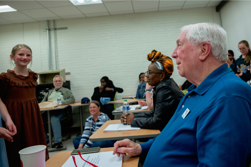An audience and panel of judges after assessing a student performance. We see one older white man with white hair and a Black woman.  The student visible is a blonde middle or high-school aged white girl wearing a long maroon dress.