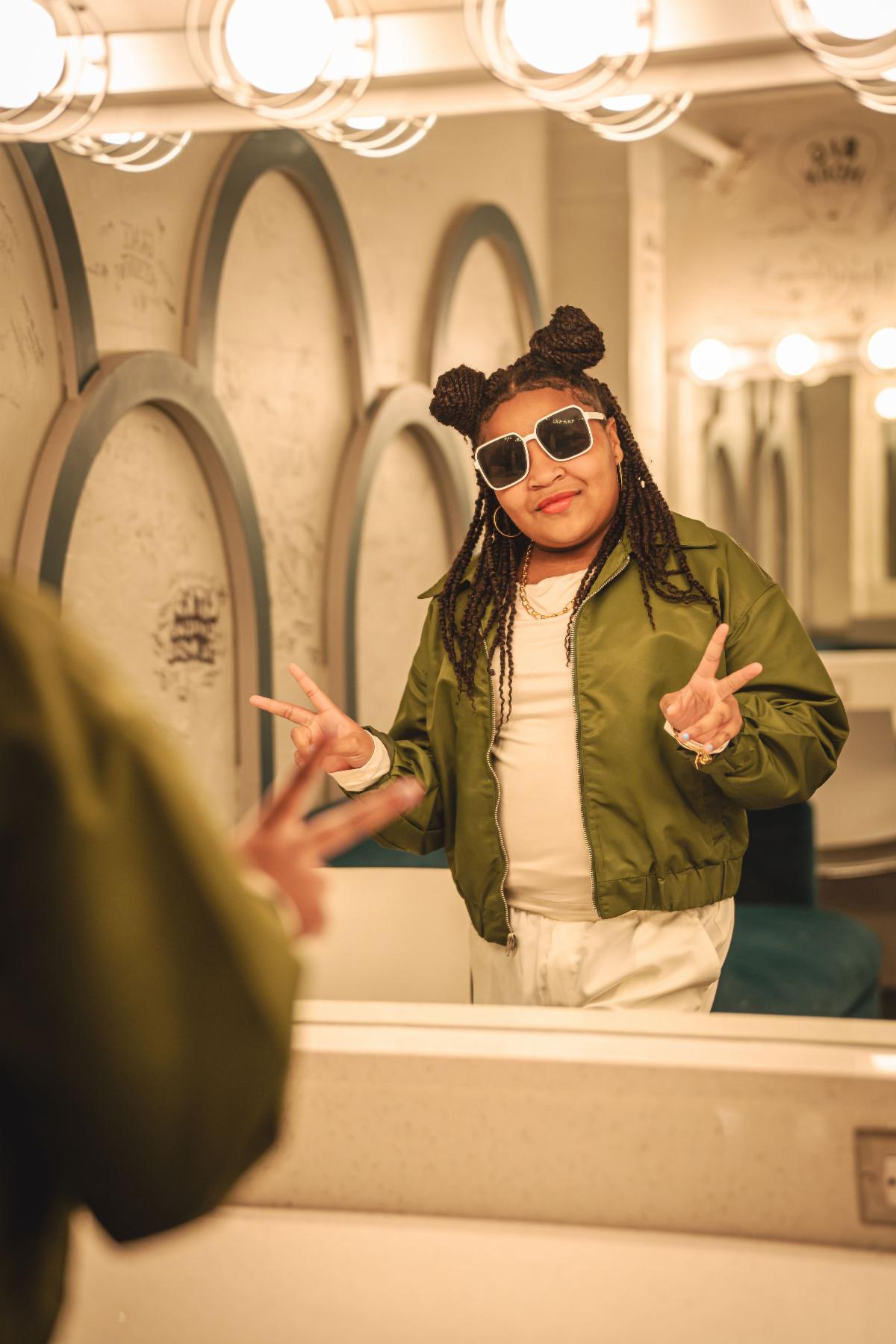 A Black child or teenage backstage before a performance makes the double peace sign in the dressing room mirror. She wears sunglasses.  Her hair in is in locs with space buns.