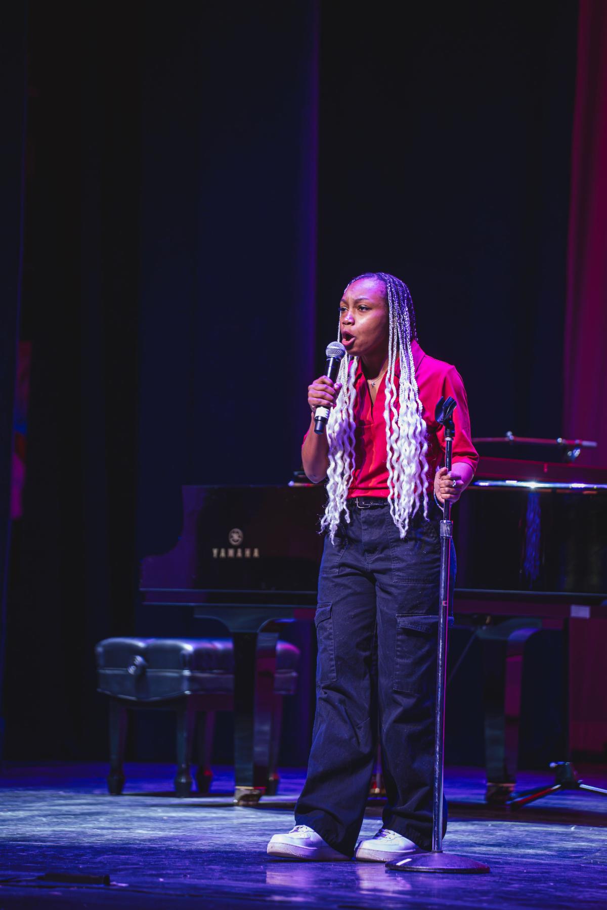 A Black teenage girl sings or speaks with a microphone. She has long hair in locs. The top is black and the bottom are light blonde or white. She wears a red button up top and black pants.
