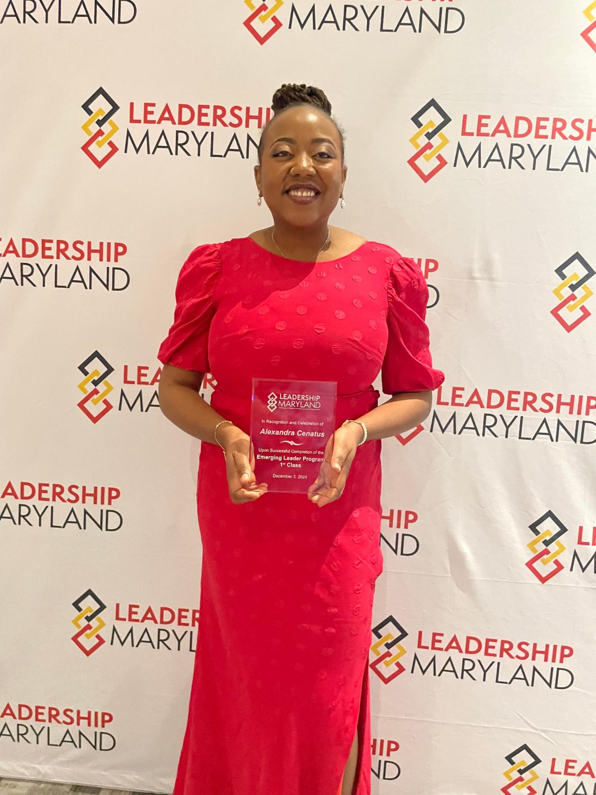Alexandra Cenatus is a Black Haitian woman who is middle aged. She holds a plaque with the Leadership Maryland logo and unreadable text. She stands in front of a step and repeat with the Leadership Maryland logo. Alexandra wears a long red dress with a high neck and three quarter sleeves. Her hair with locs is in an updo.