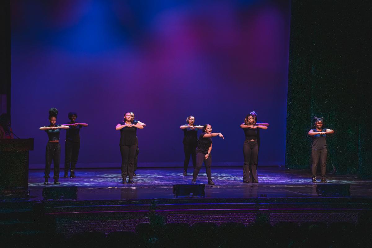 A group of ten Black teen girls performing either a dance or physical performance art. They all wear black shirts and pants. The girl in the front points down.