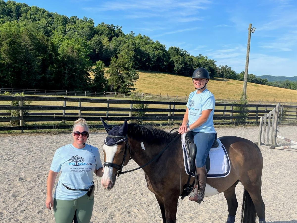 A person stands next to another person, who is on horseback. Both wear Giving Day shirts. 