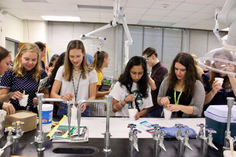 A row a female students stand as a table in an LBC lab. Their heads are bent over their tasks and they are each stirring something in a styrofoam cup.