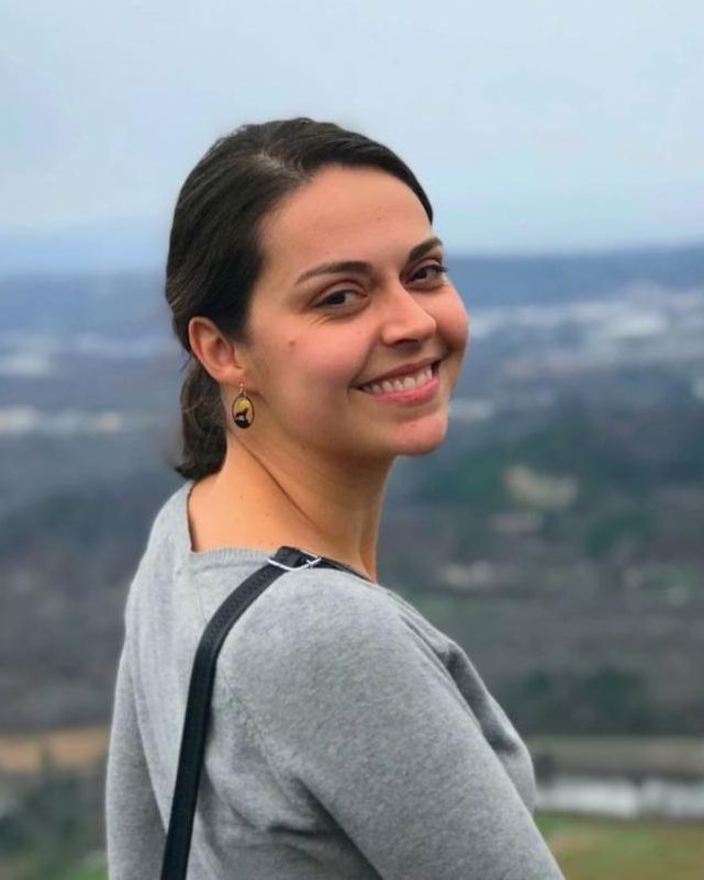 Amanda Domingues has dark hair and a gray sweater. There's scenery in the background