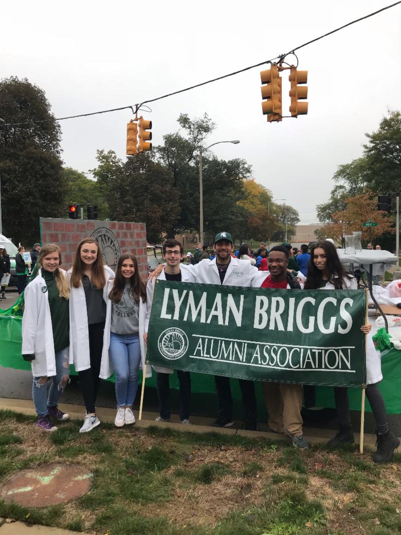 A group of students in lab coats stand in front of the float holding a banner that reads "Lyman Briggs Alumni Association."