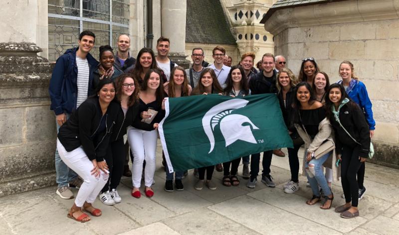 Dr. Ward and Dr. Parks and a group of students hold up a Spartan flag outside of a building in Paris. 